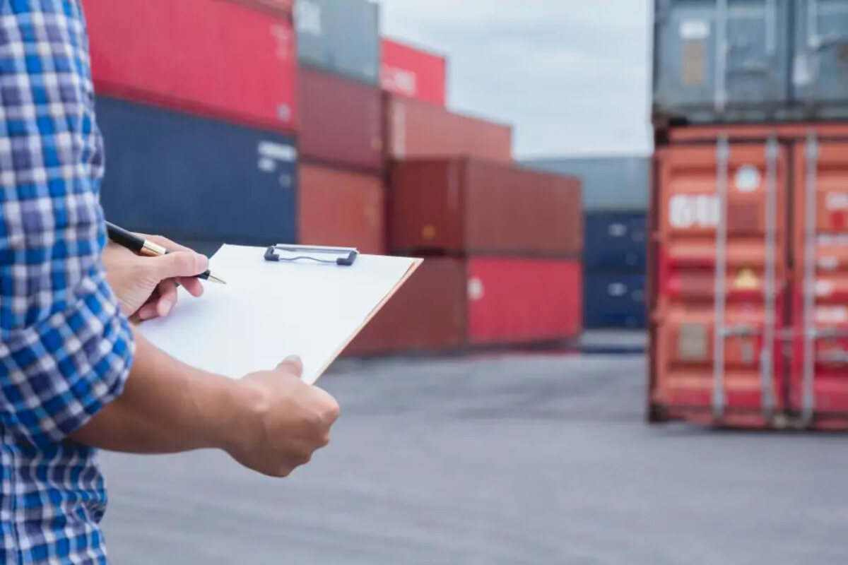 Man writing on clipboard standing in front of shipping containers