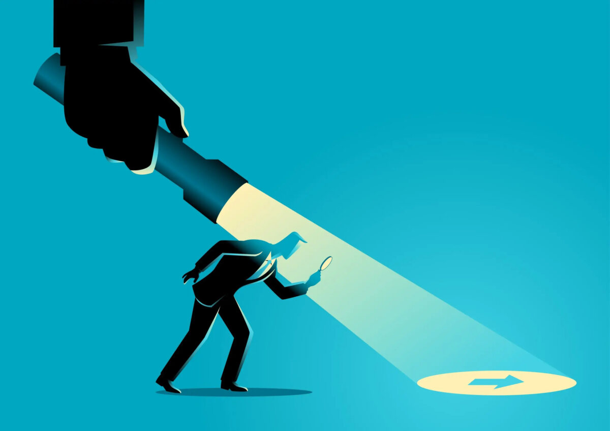 Cartoon of a businessman looking through a magnifying glass at an arrow on the floor that is illuminated by a giant torch held from above by a giant hand