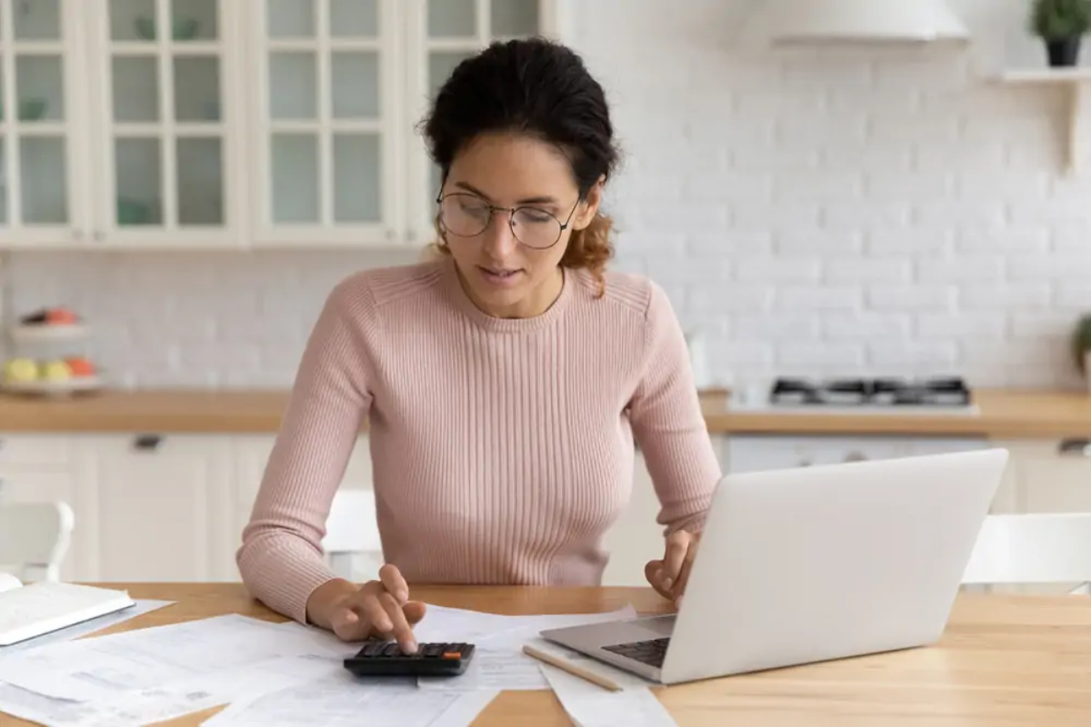 Businesswoman at kitchen table using laptop with one hand and calculator with the other