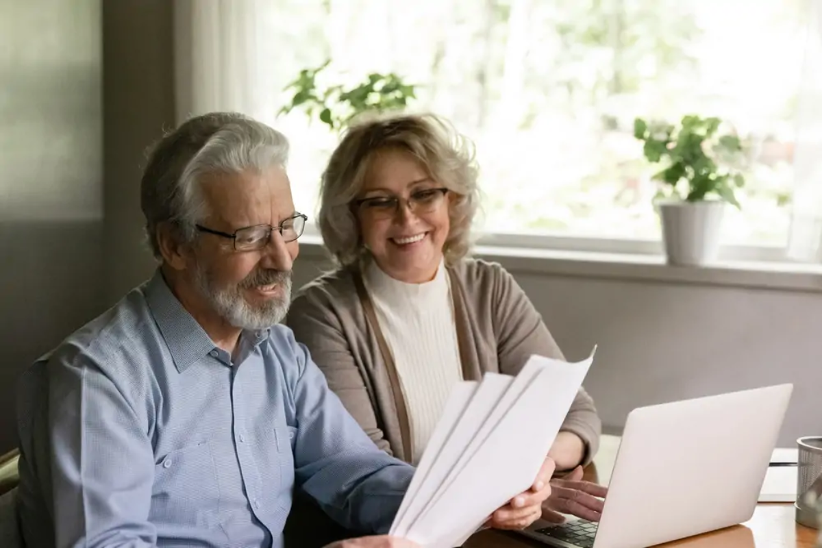 Elderly couple at table with open laptop holding and reading National Insurance and State Pension while smiling