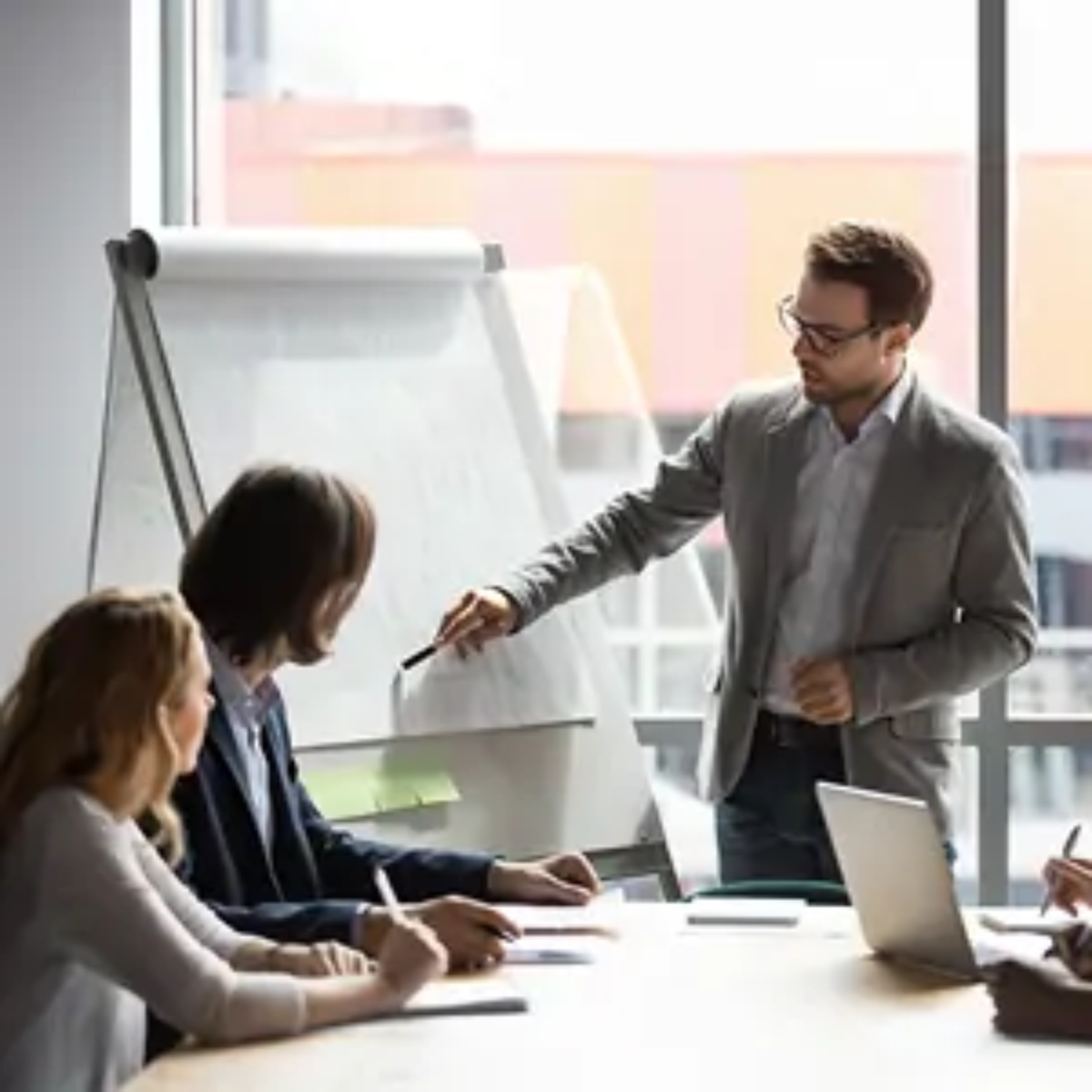 Businessman standing pointing at flip chart with pen and talking to colleagues sitting down in business finances meeting