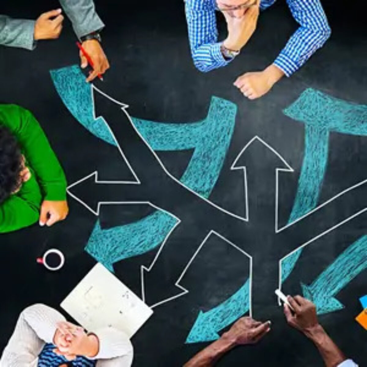 Businesspeople in meeting sitting around chalk board-style desk with arrows pointing in different directions drawn on it