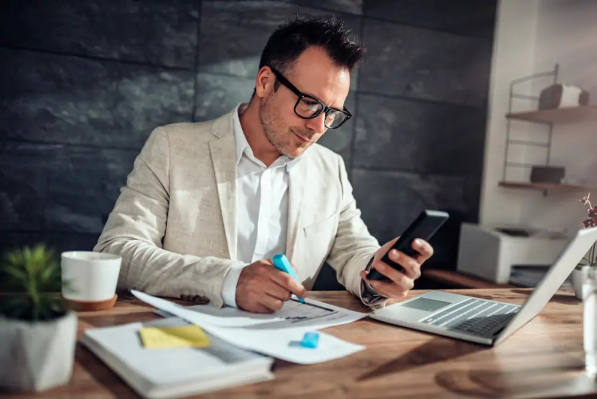 Businessman at desk using smartphone in one hand and holding a highlighter in the other over management accounts document