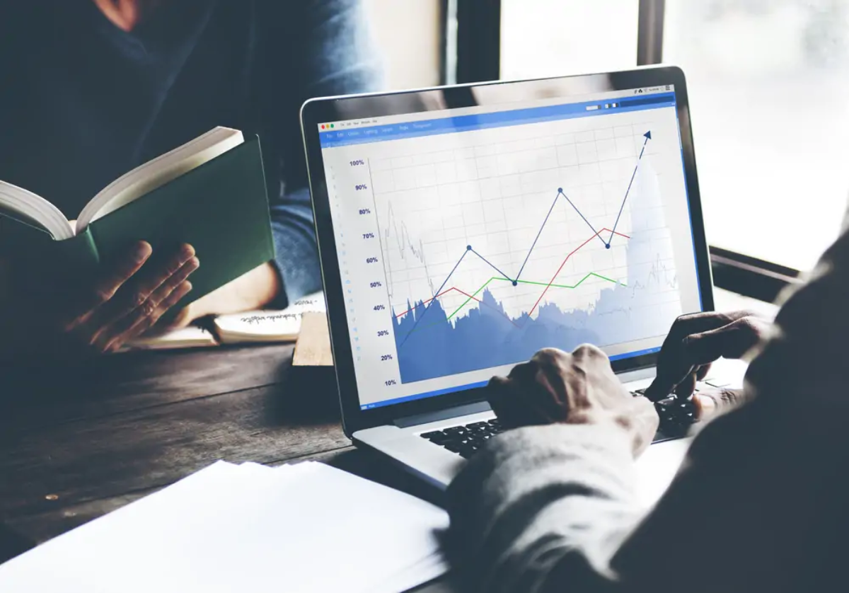 Businessperson at table using laptop with business growth graph displayed on screen opposite another businessperson reading a book