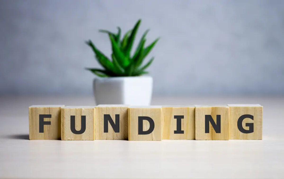 Wooden blocks with letters printed on lined up to spell 'funding' in front of a plant pot