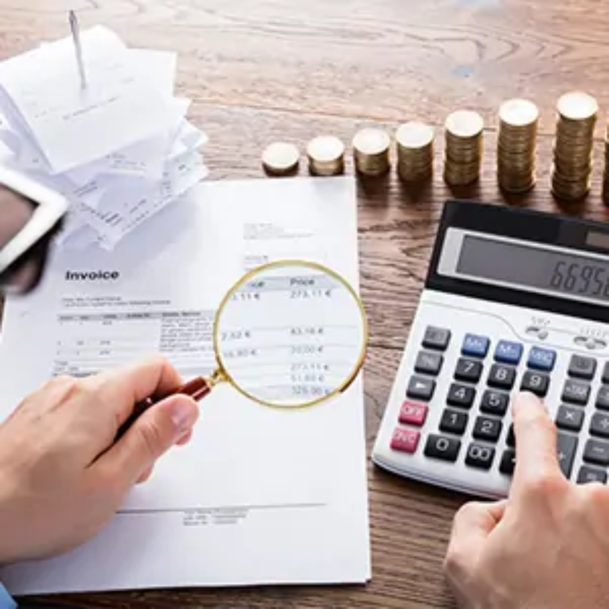 Person carrying out tax investigation at desk with one hand using calculator and the other holding magnifying glass over invoice