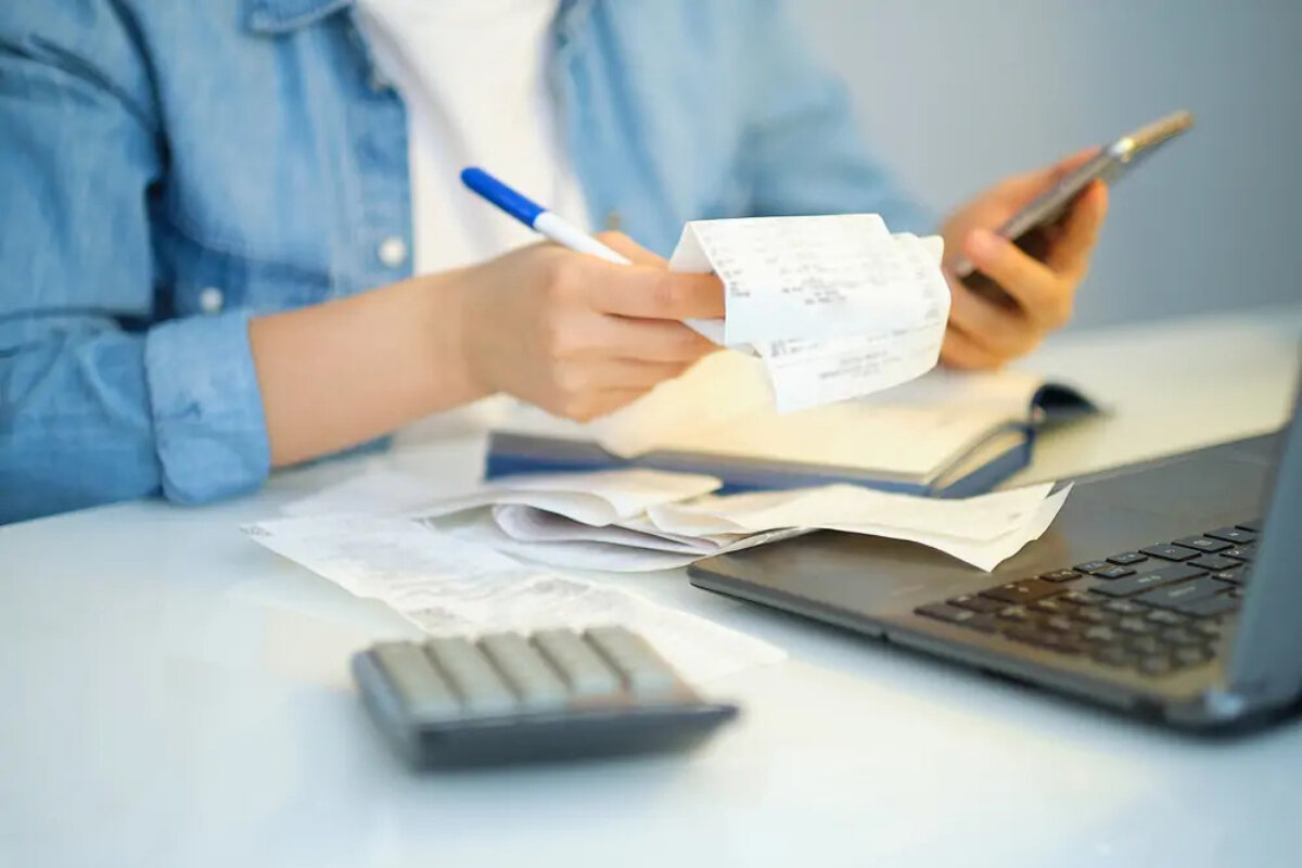 Businessperson holding smartphone in one hand and receipts and a pen in the other over an open account book and laptop