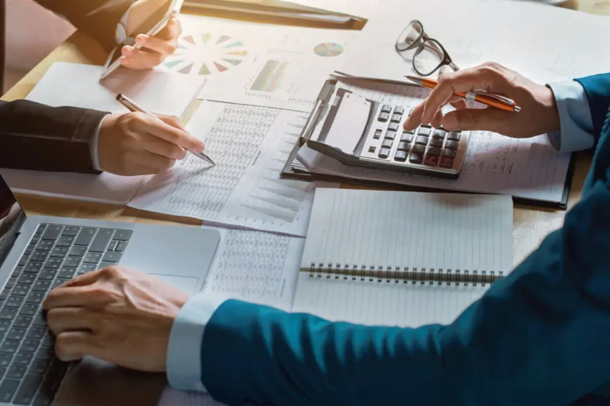 Two businesspeople sitting opposite each other at desk, one using a laptop and calculator and the other holding a smartphone and writing on a document