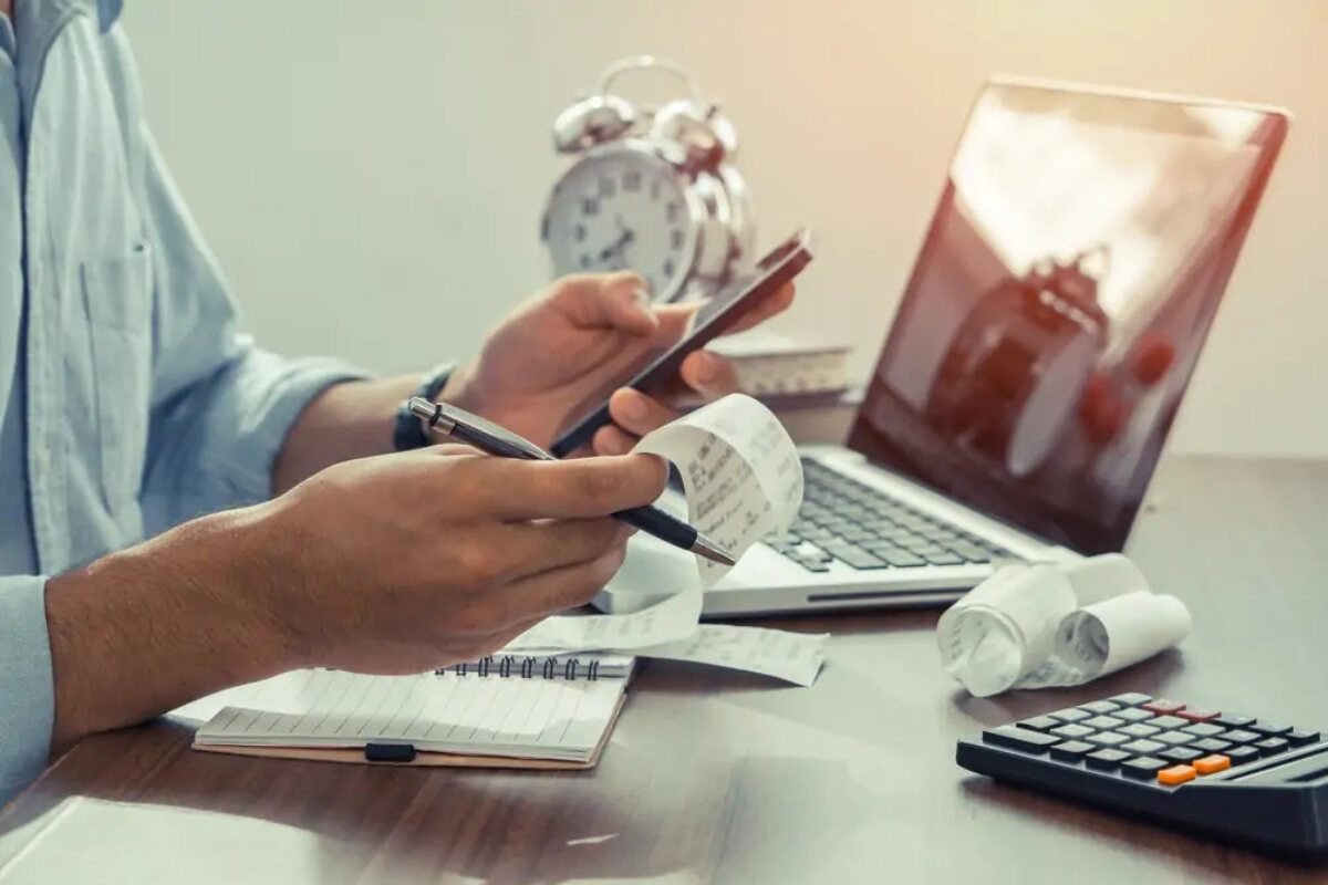 Businessman at desk holding smartphone in one hand and pen and receipts in the other