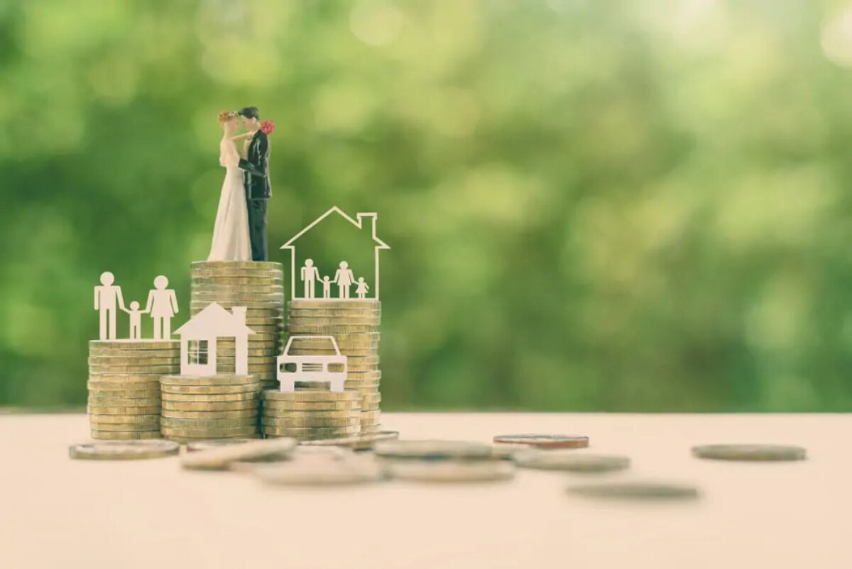 Five stacks of coins of varying heights, one with a paper cutout of a car on top, one a house, one two parents and a child, one a house with a family inside, and one a wedding cake topper