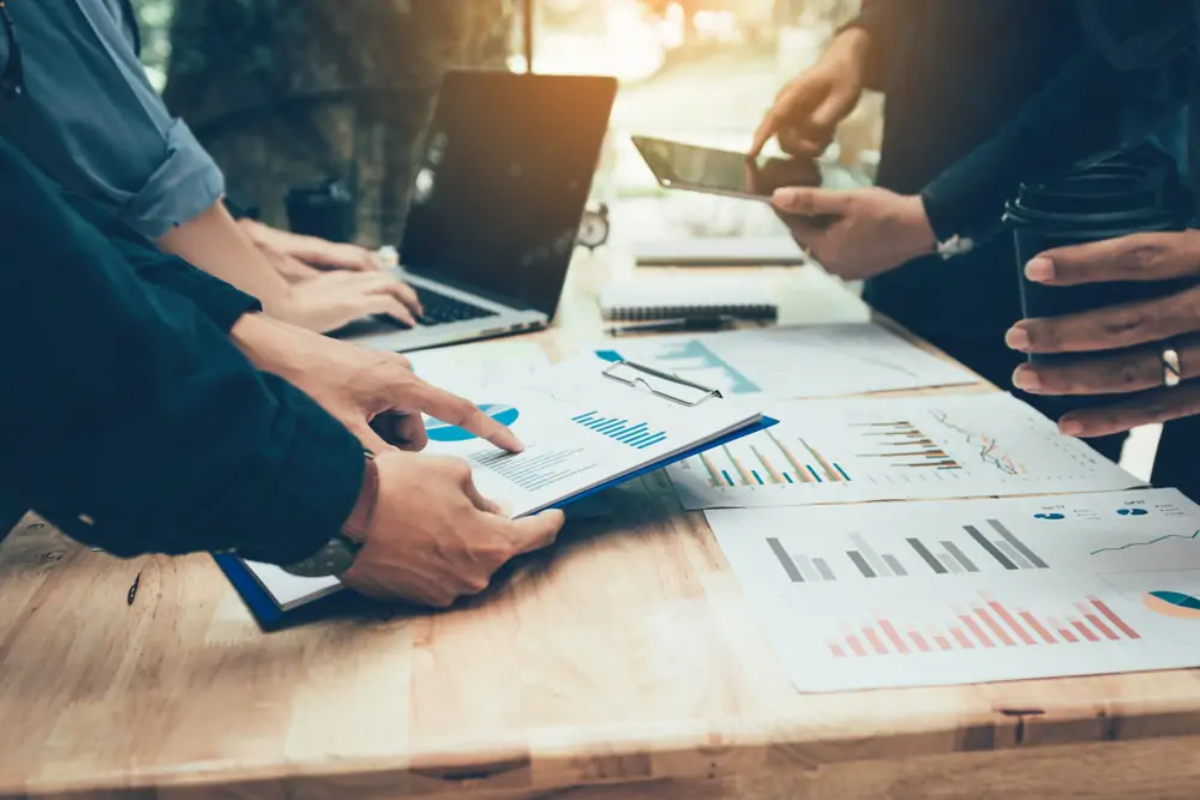 Businesspeople standing around table with laptop, tablet and pointing at printed graphs