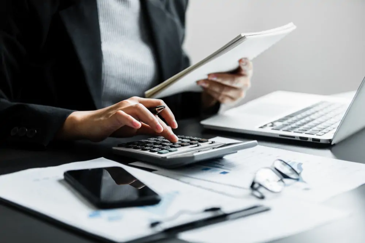 Businesswoman at desk holding notebook in one hand using calculator with the other making Capital Gains Tax calculations