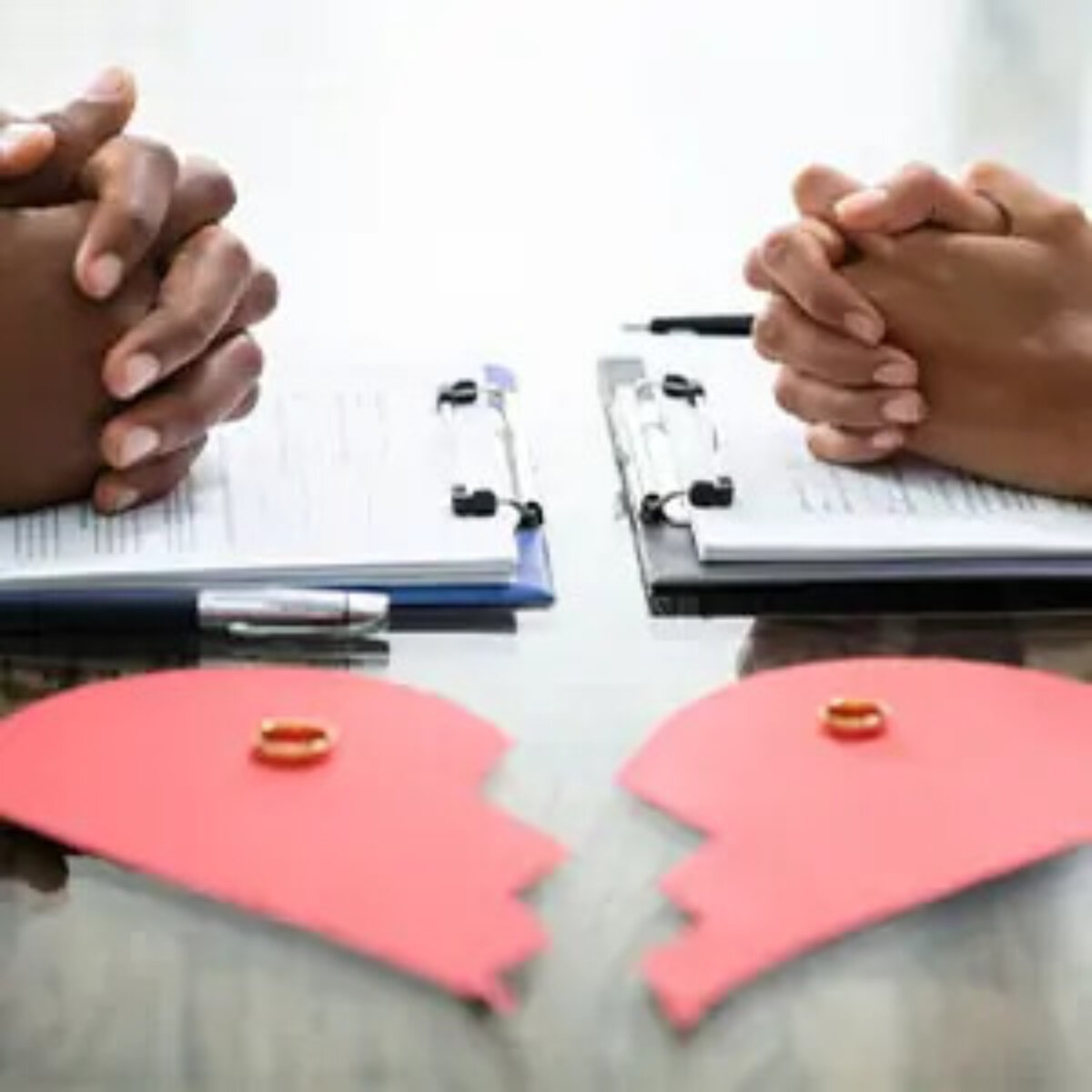 A separating couple opposite each other with their hands clasped over separate clipboards and a broken paper heart in front with wedding rings on top