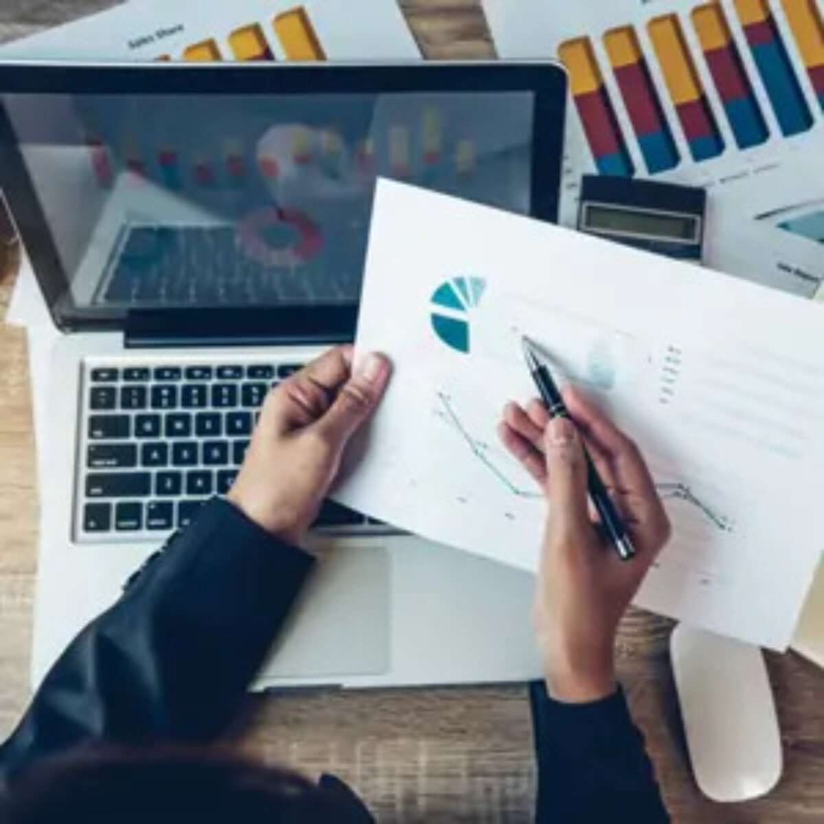 Businessperson holding pen over graph document above open laptop