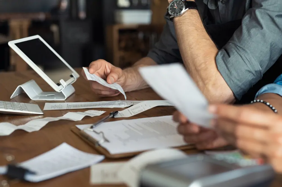 Two business owners at table holding receipts and using tablet to calculate payments