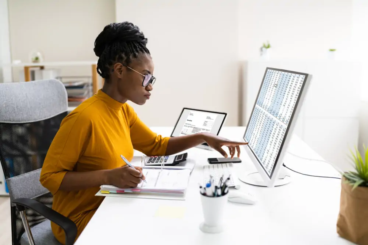 Businesswoman at desk pointing at computer screen and writing on document in ring binder