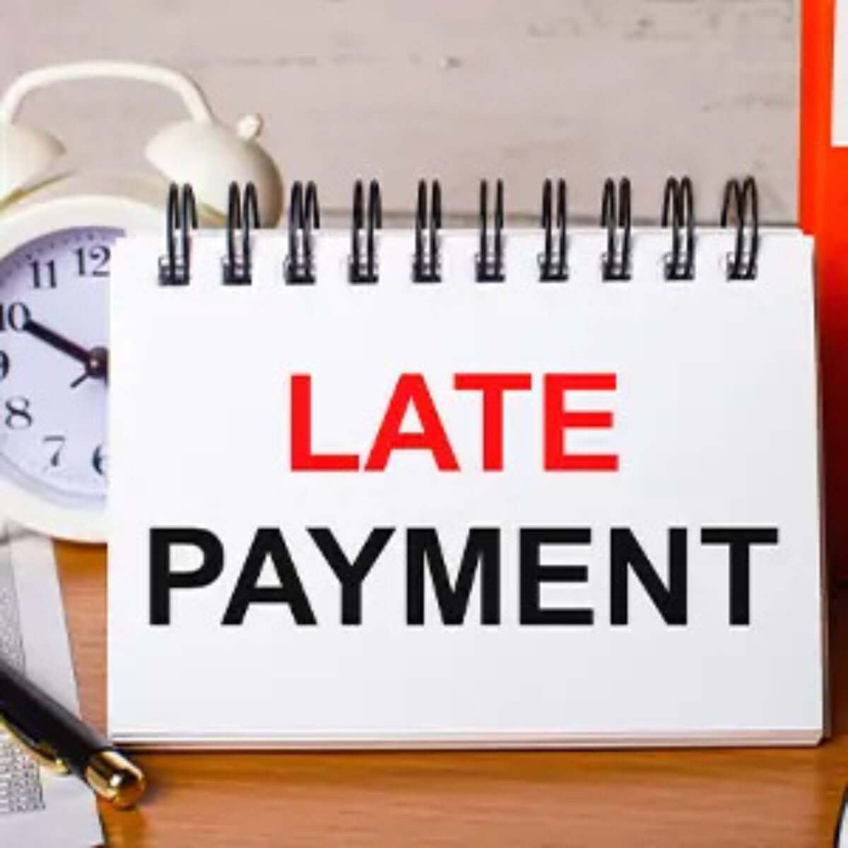 Upright open ring-bound notebook with 'late payment' on page on desk in front of alarm clock