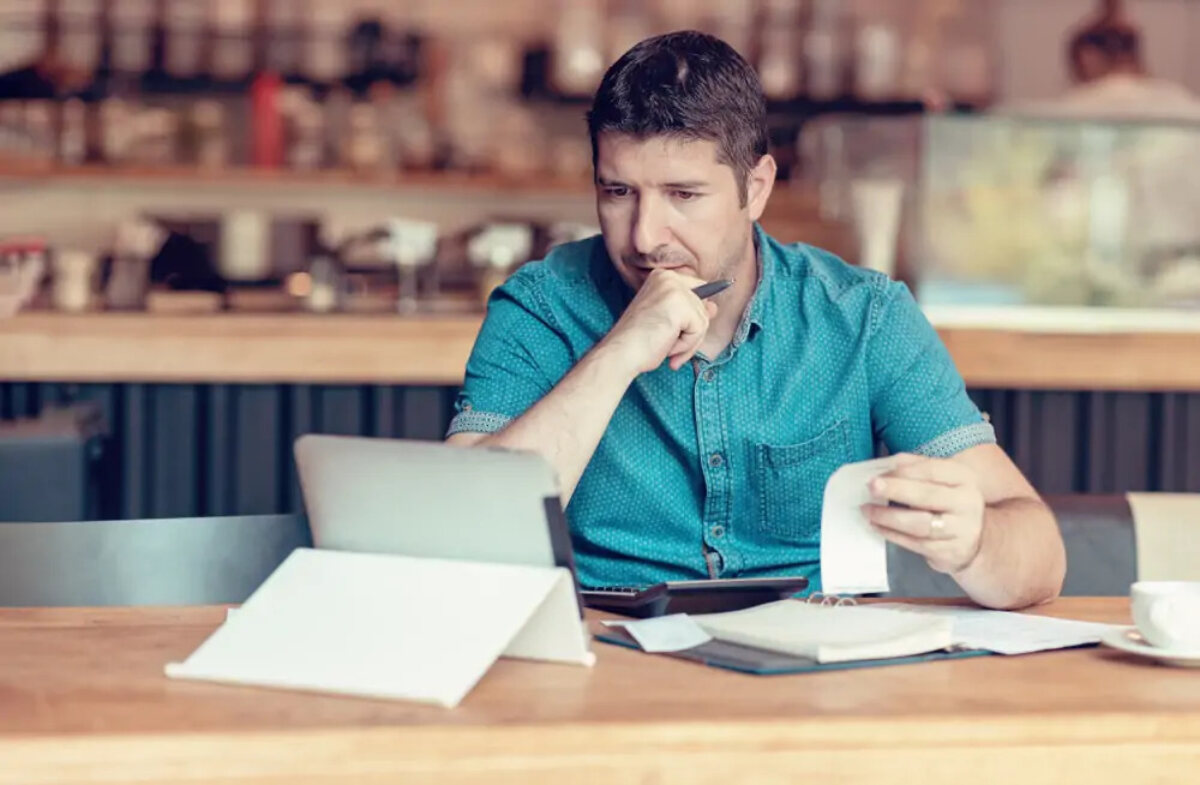 Cafe owner making calculations with receipts, tablet and calculator at table in cafe
