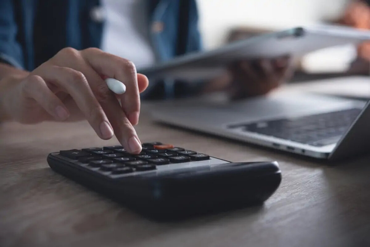 Businessperson calculating tax at desk while holding tablet ad using calculator next to open laptop