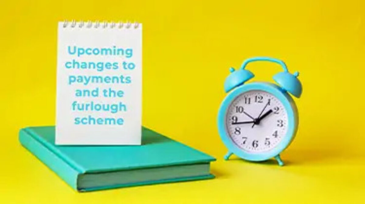 Alarm clock next to closed book with open notebook on top with page displaying 'upcoming changes to payments and the furlough scheme'