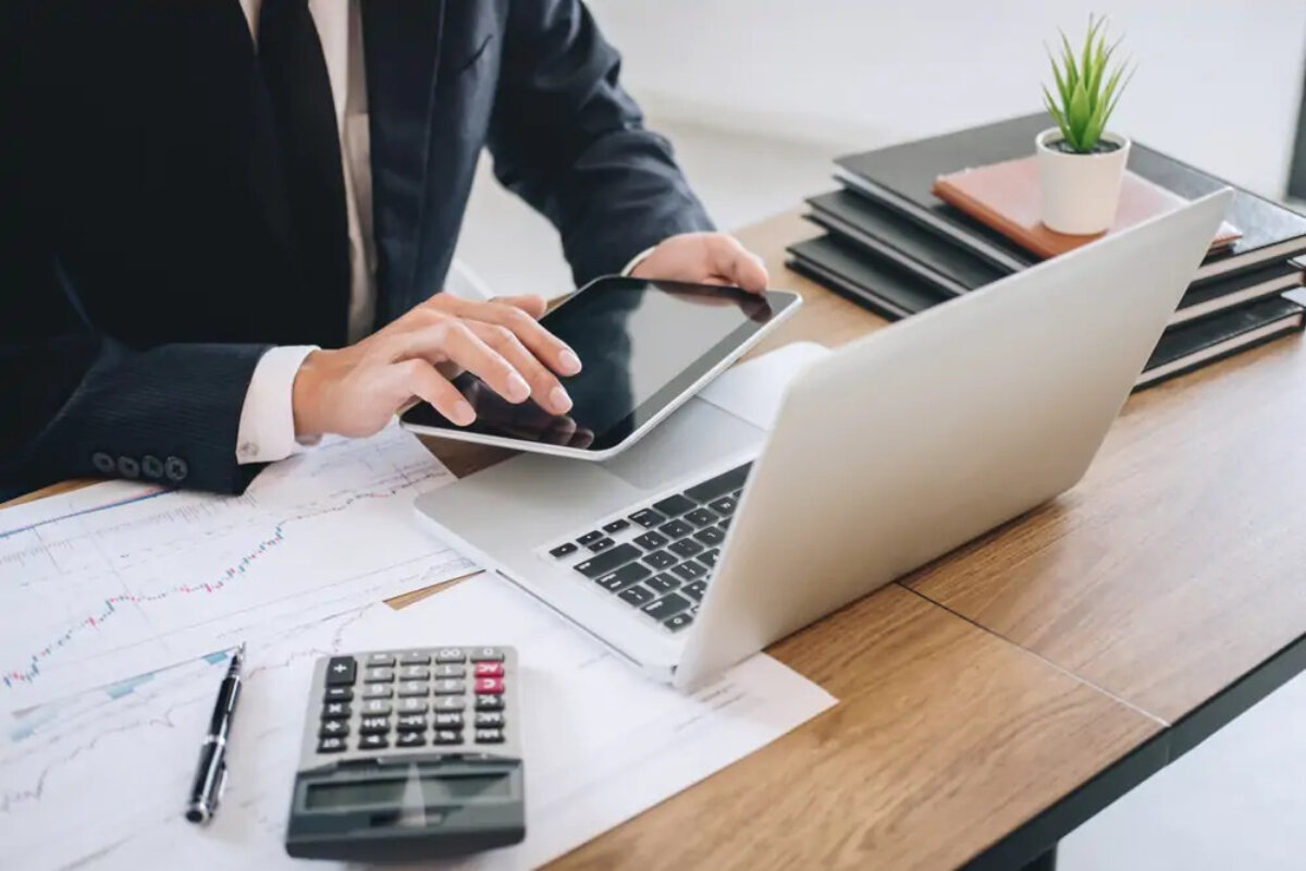 Businessman using tablet in front of open laptop next to calculator and graph documents