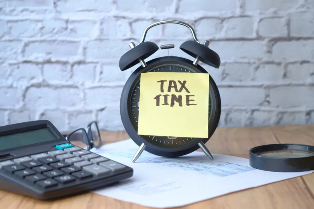 Alarm clock on desk on top of HMRC document and next to calculator and magnifying glass with post-it note reading 'tax time' over face