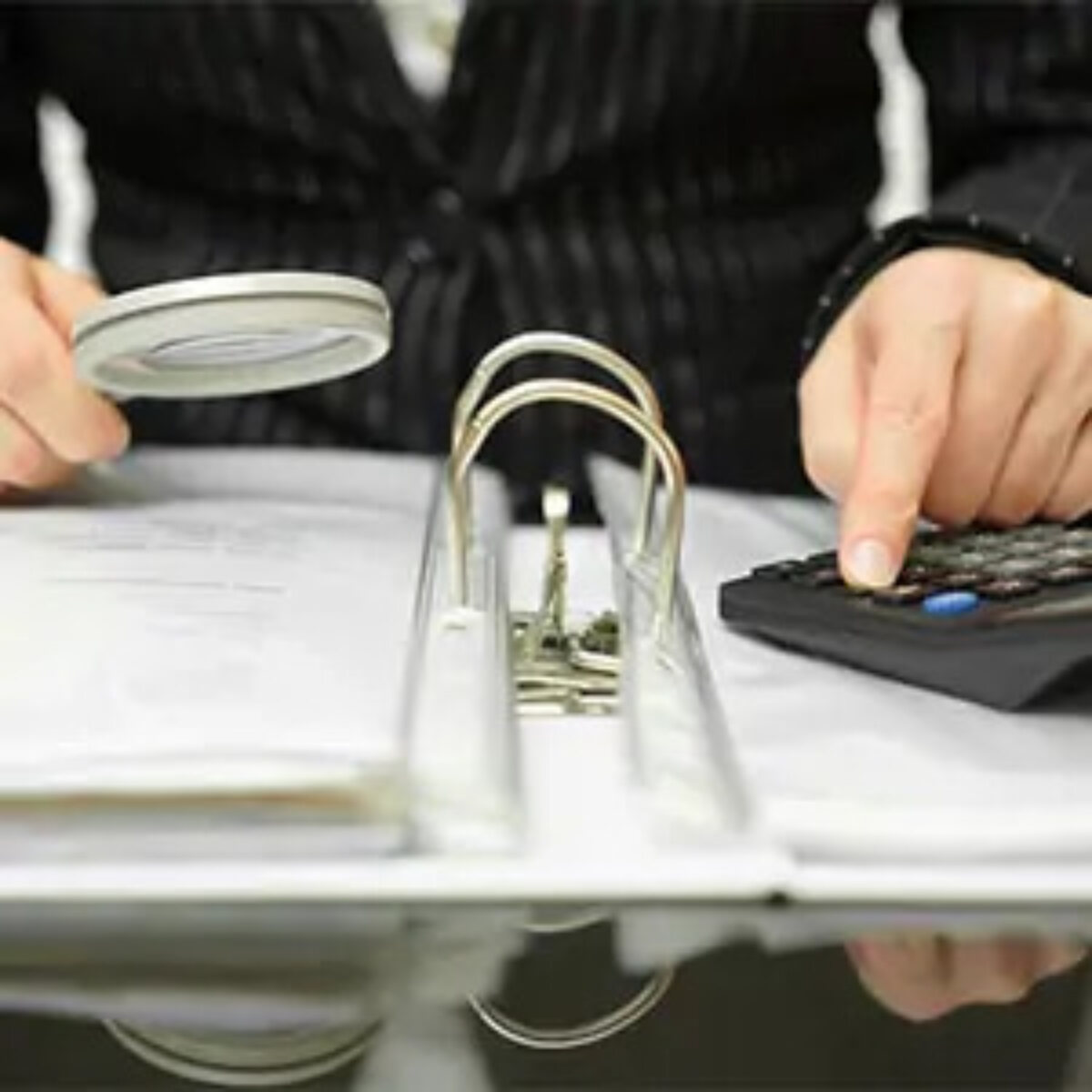 Investigator holding magnifying glass in one hand and using calculator with the other on top of an open ring binder