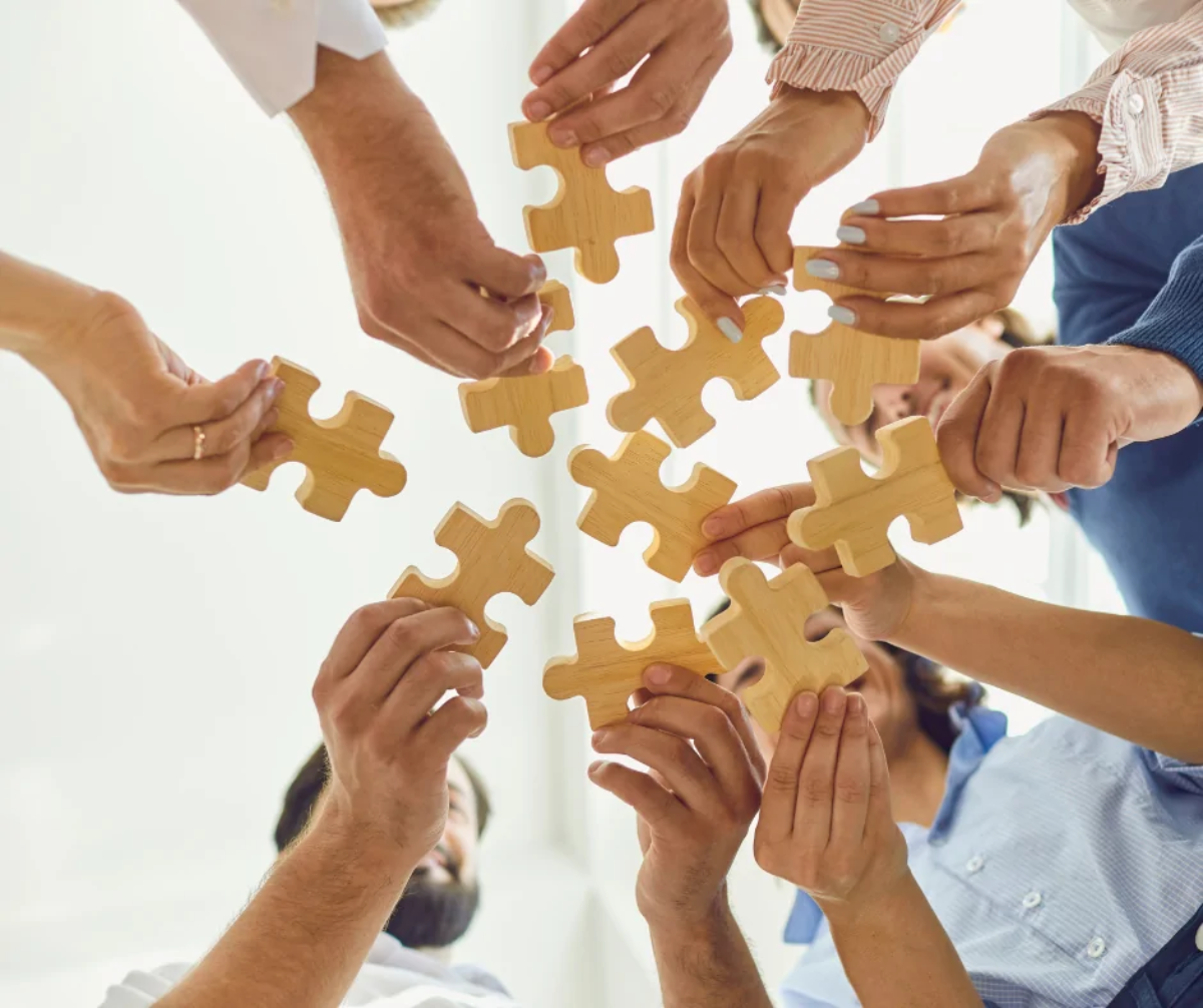 Ten hands in a circle holding a wooden jigsaw piece each in the middle