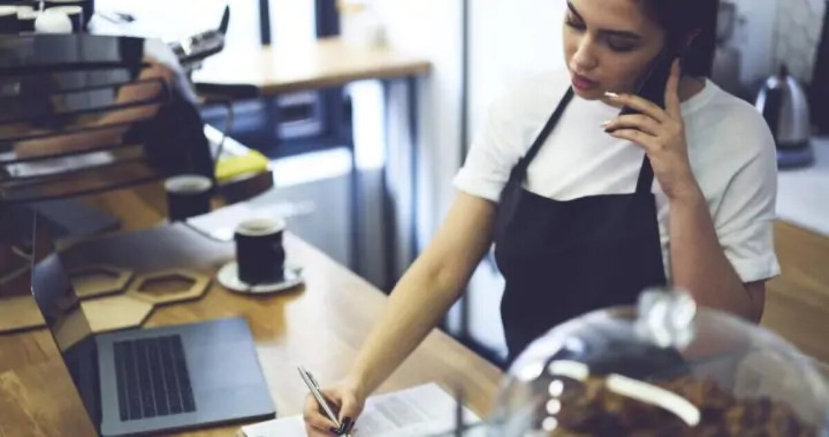Barista behind coffee bar on the phone noting things down on a document next to an open laptop