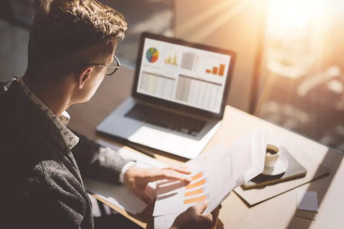 Businessman at desk looking at laptop screen and holding document displaying annual accounts