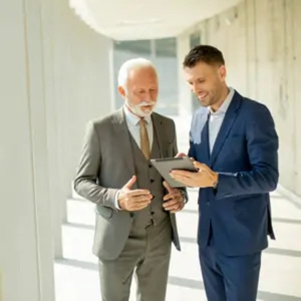 Younger businessman standing talking to elderly businessman and holding tablet that they are both looking at