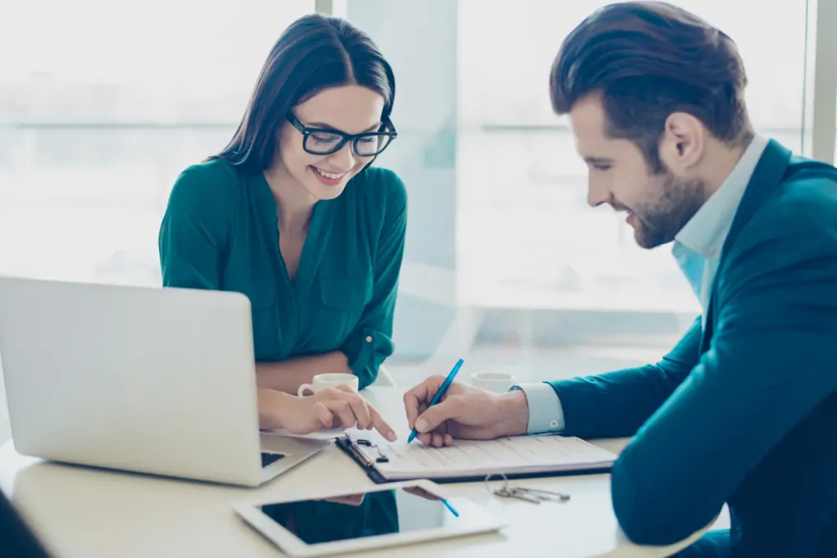 Two businesspeople sitting at desk looking and pointing at document on clipboard