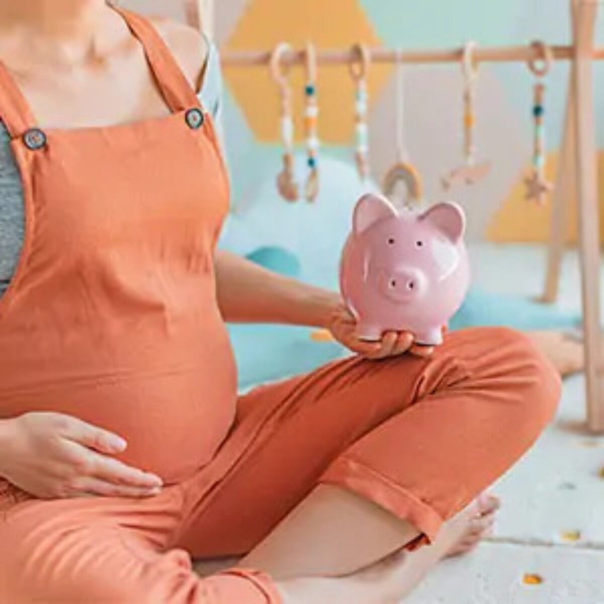 Pregnant woman sitting cross-legged on floor of nursery holding stomach with one hand and a piggy bank with the other