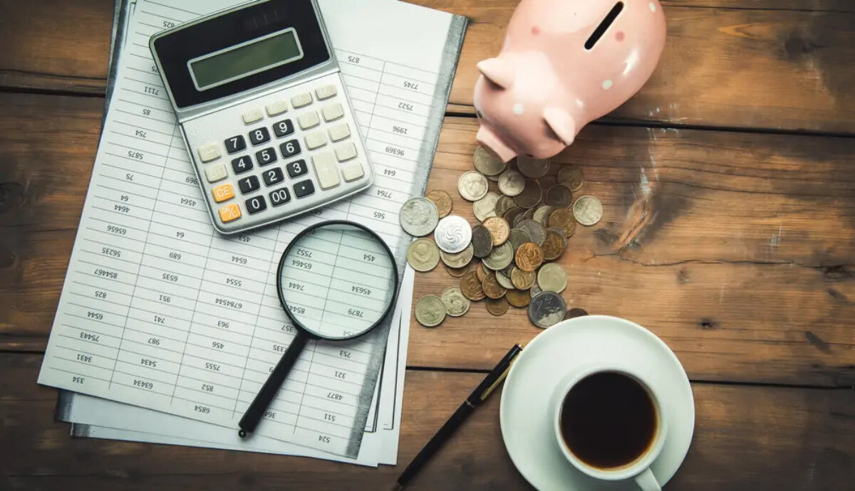 Calculator and magnifying glass on top of document, next to piggybank, pile of coins, and cup of coffee on wooden table