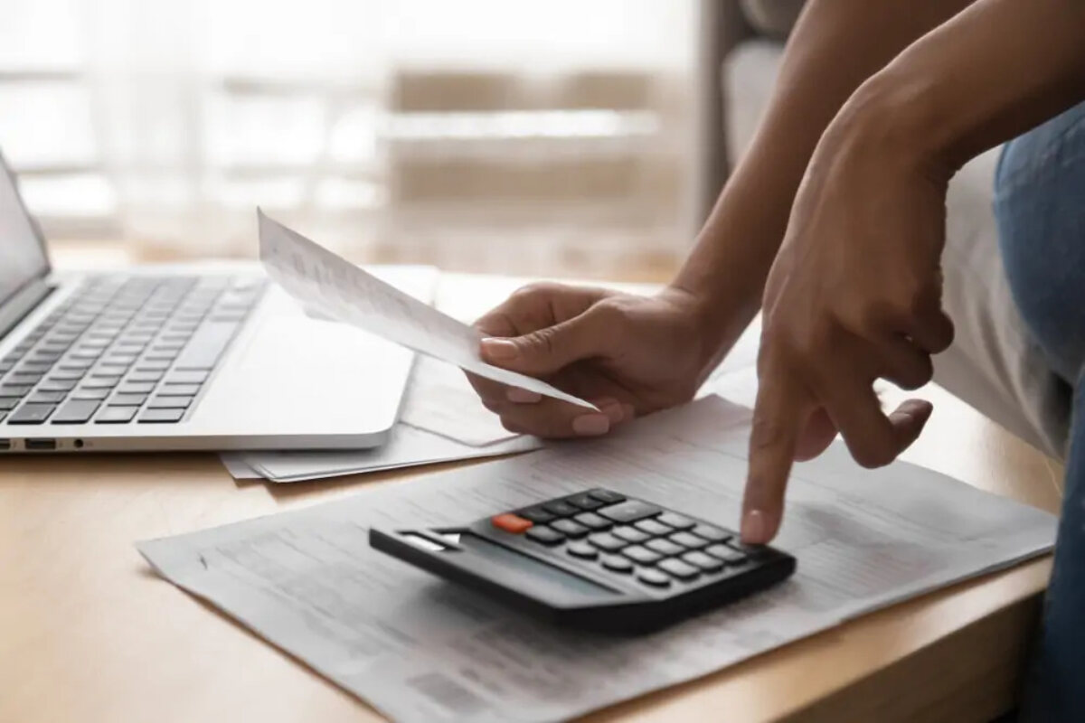 Woman holding paper in one hand and using calculator with the other next to open laptop