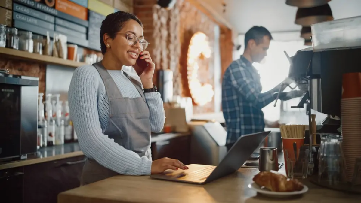 Cafe owner taking phone call and using laptop behind coffee bar