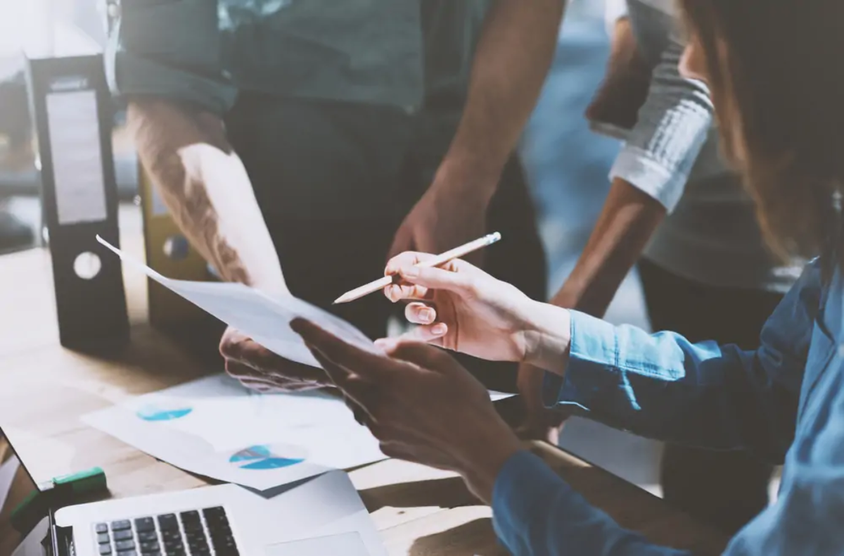 Businesswoman sitting at desk holding up document and pointing at it with pencil explaining to colleagues standing next to her how to read a set of management accounts