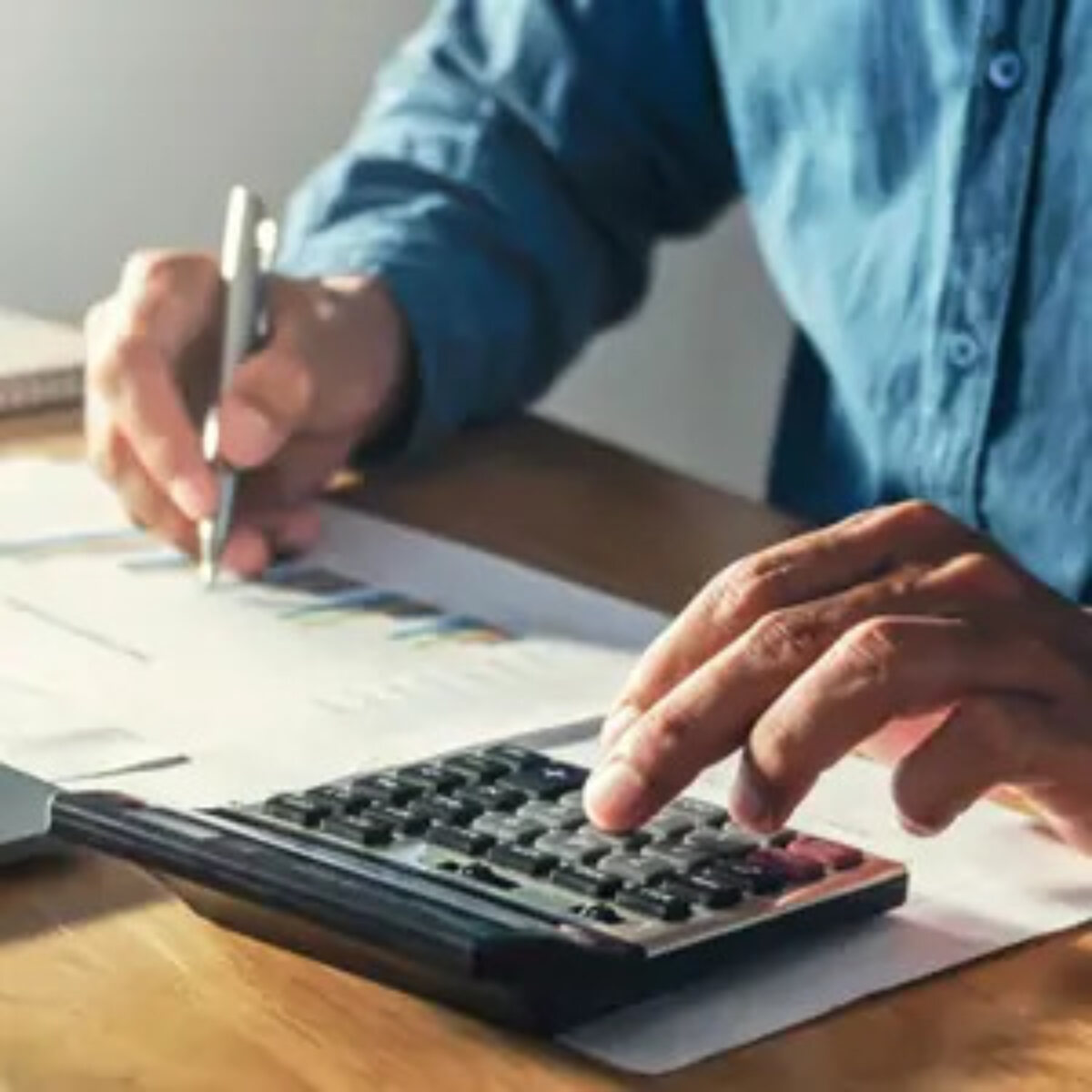 Businessman using calculator with one hand and holding pen to write on documents in the other