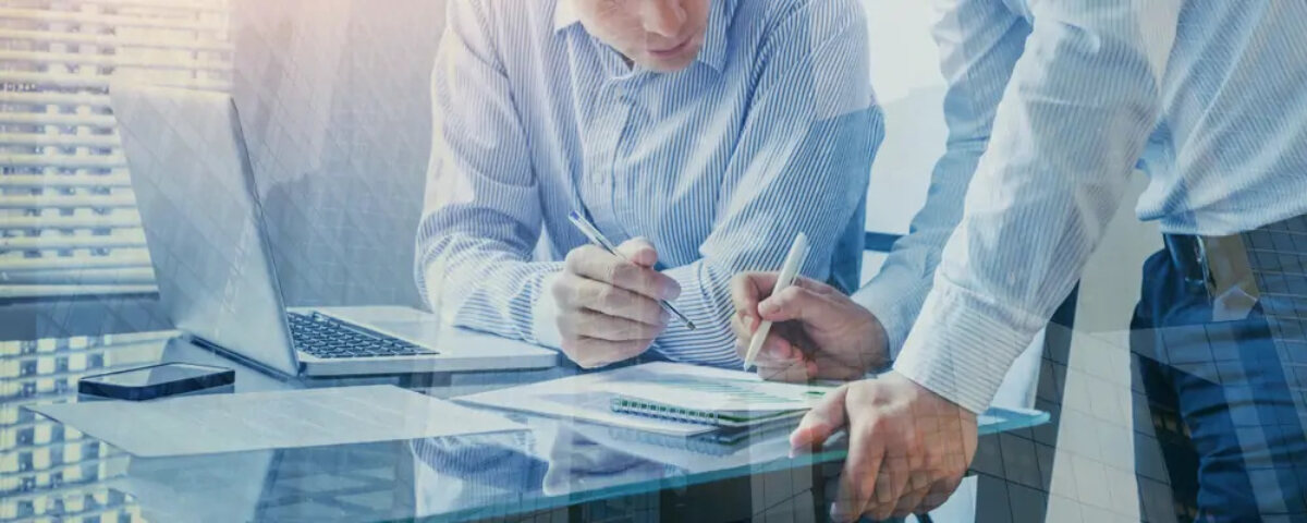 Two businessmen at desk with open laptop looking at and writing on Corporation Tax document