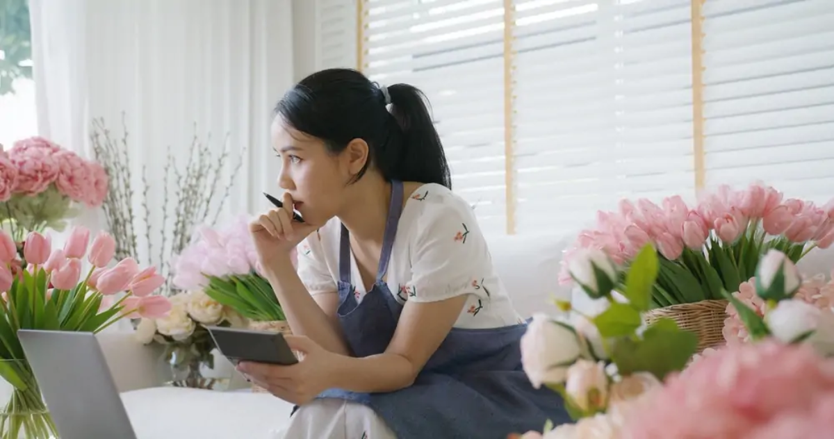 Florist sitting in front of laptop in shop with calculator in hand making cash flow calculations
