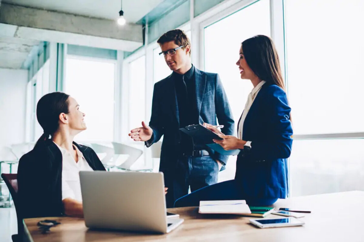 Businesswoman sitting at desk talking to two colleagues standing next to her
