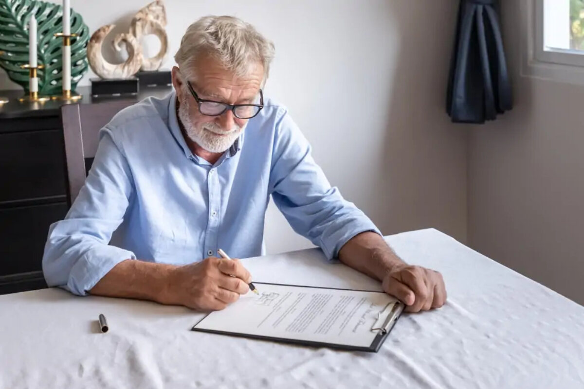 Elderly man at table signing form on clipboard