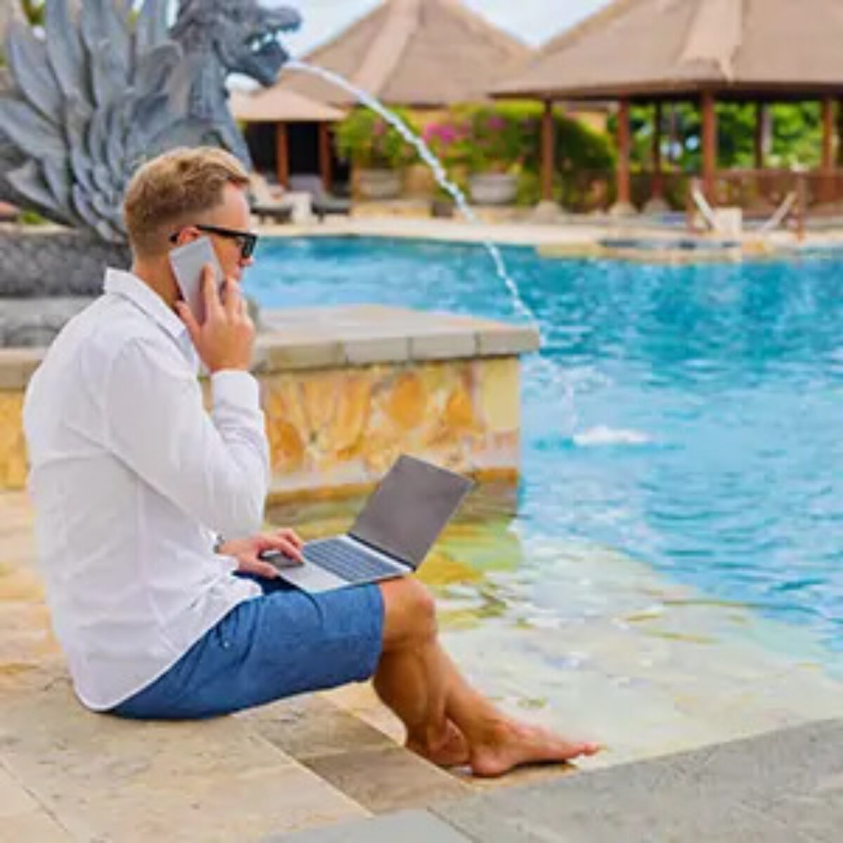 Businessman sitting on steps of outdoor swimming pool with laptop on lap and talking on phone