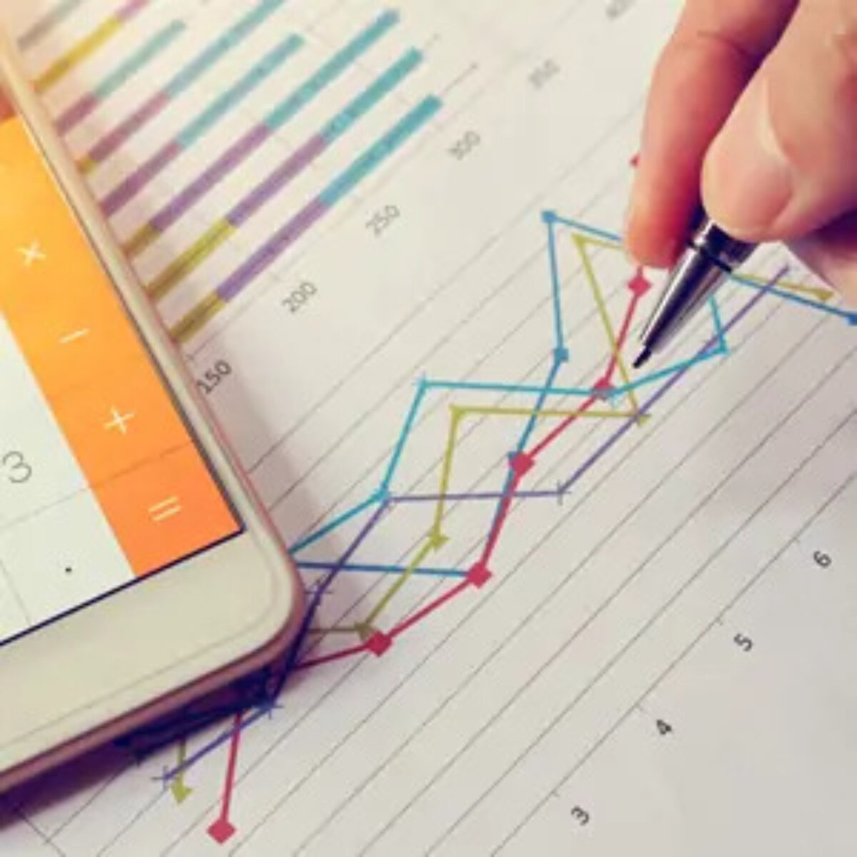 Hand plotting line graph on paper with pen and calculator app open on smartphone