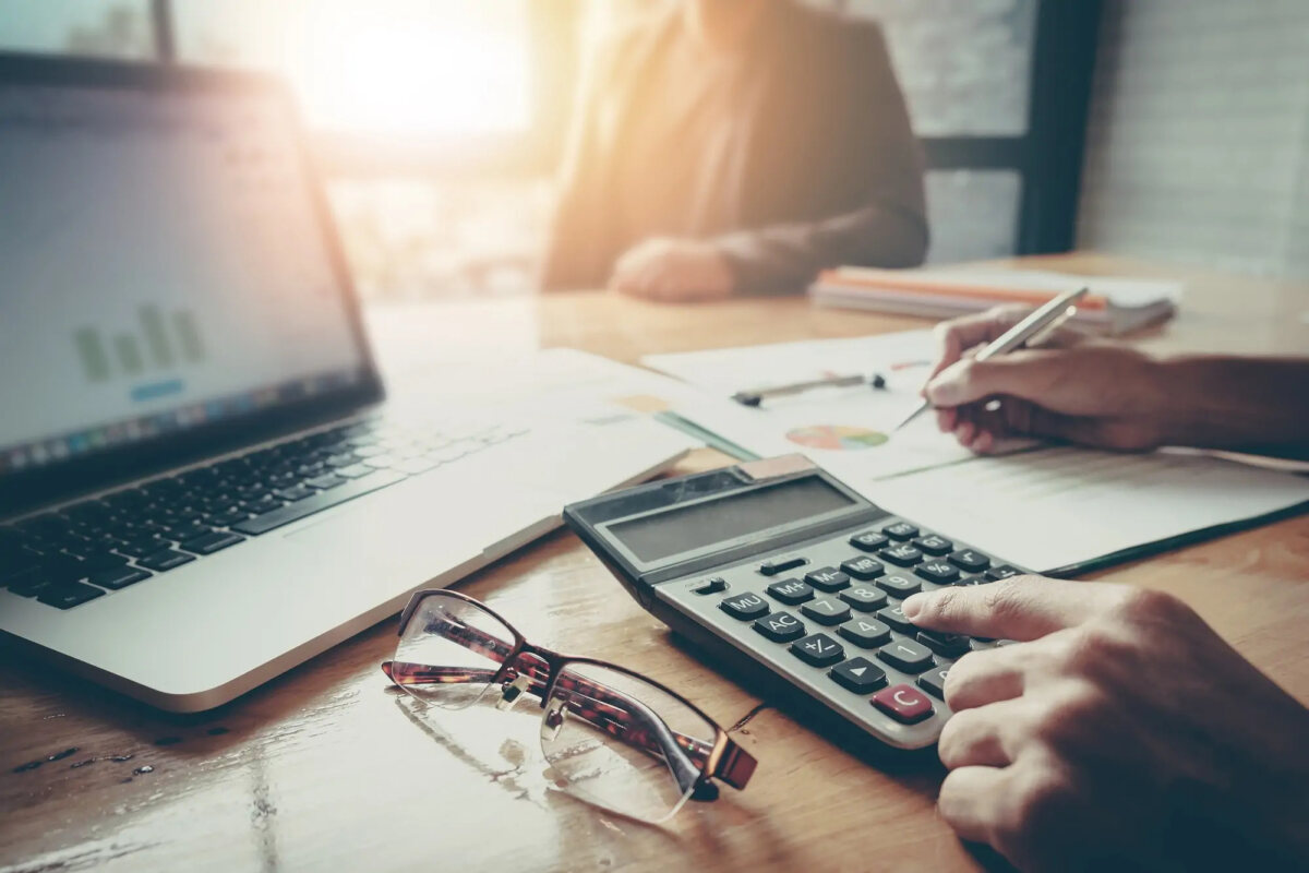 Businessperson at desk using calculator with one hand and writing on document with the other