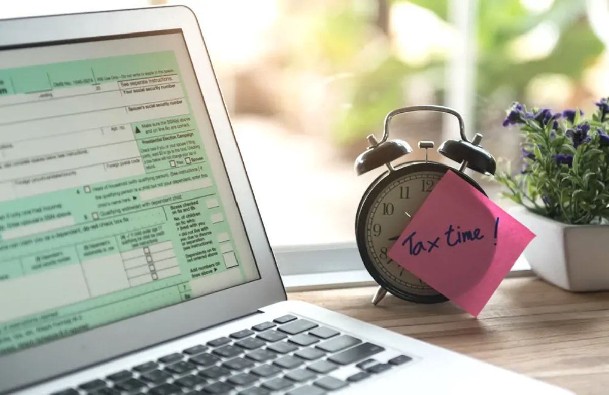 Alarm clock with post-it note on face saying 'tax time!' next to laptop with screen displaying Self Assessment form