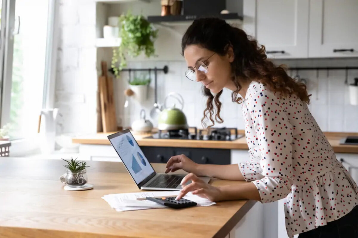 Businesswoman standing in kitchen using calculator and laptop on island to calculate year-end taxes