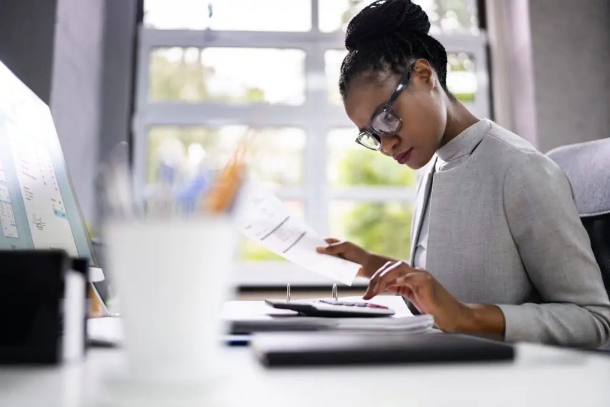 Businesswoman at desk holding accounting document in one hand and using calculator with the other