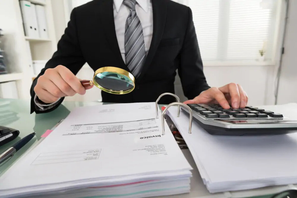 Businessman at desk using calculator with one hand and holding magnifying glass over invoice document in ring binder in the other to investigate business fraud