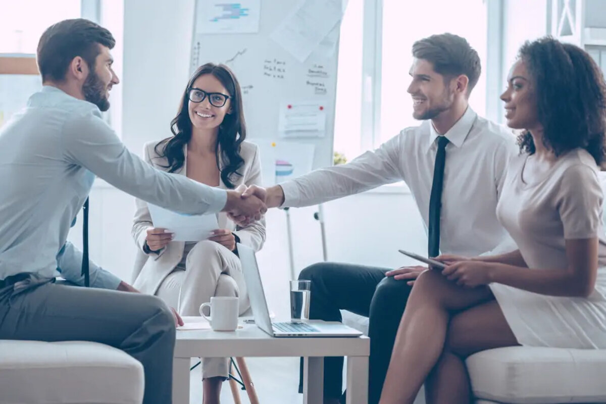 Four businesspeople sitting around coffee able at meeting; the two businessmen are shaking hands over the table